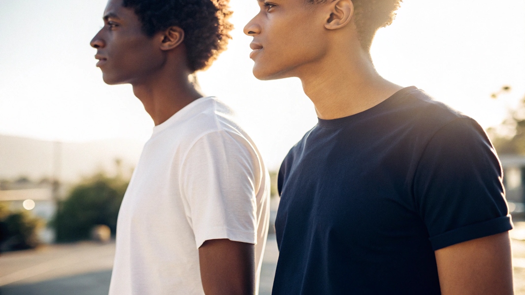 A side-by-side image of two people wearing T-shirts: one in a light-colored (white or pastel) T-shirt and the other in a dark-colored (black or deep blue) T-shirt, both standing under bright sunlight. The person in the light-colored tee appears cooler and more refreshed, while the dark-colored tee wearer looks warmer. The focus is on subtle expressions, clothing texture, and the outdoor setting with soft lighting, highlighting the visual temperature contrast between the two outfits.