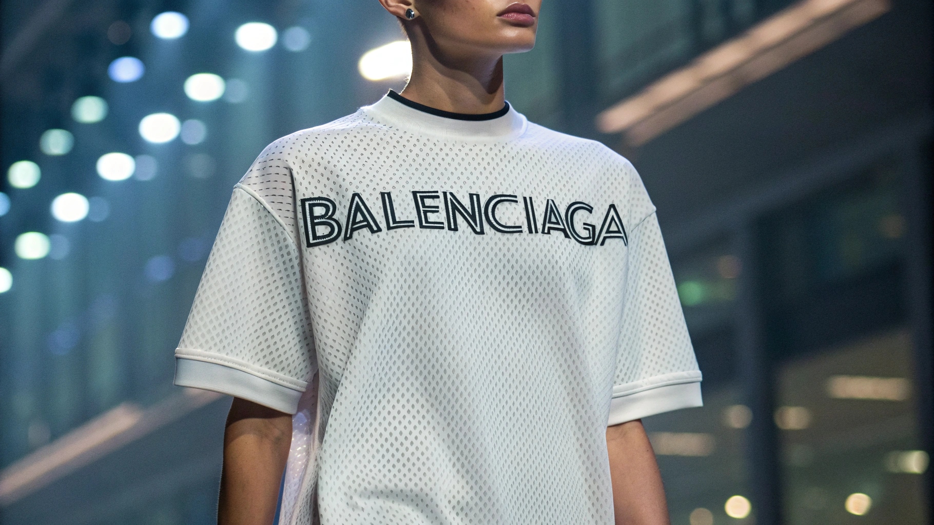 Fashion-forward model wearing a Balenciaga logo T-shirt with a bold oversized logo on the chest, featuring a minimalistic design and subtle embossed print or 3D effect. Styled in a futuristic urban backdrop, the shirt highlights high-quality fabric texture and experimental font design or color contrast. Soft lighting enhances the cutting-edge fashion vibe, showcasing a modern streetwear aesthetic.